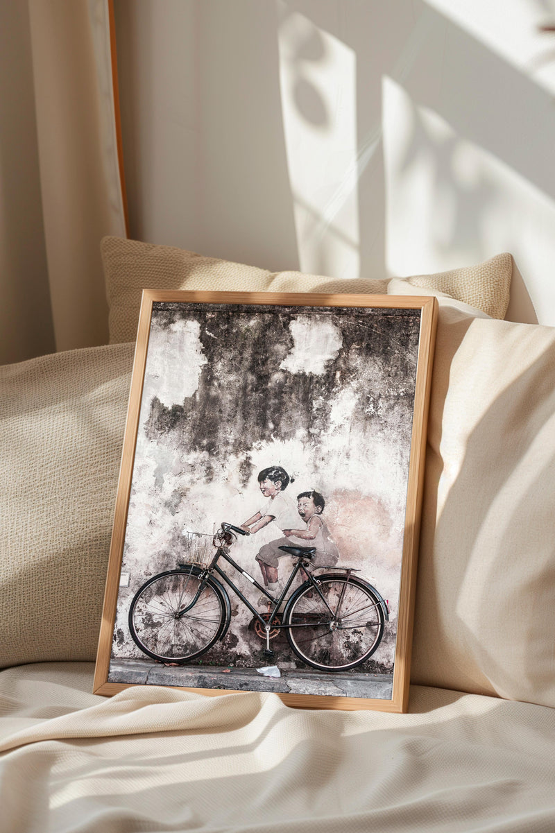 A framed black and white photograph of two children riding a bicycle is displayed on a bed with a beige pillow.