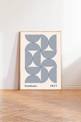 A framed poster with a geometric design featuring concentric circles in shades of blue and white, standing on a wooden floor against a white wall.