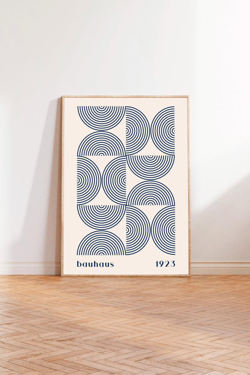 A framed poster with a geometric design featuring concentric circles in shades of blue and white, standing on a wooden floor against a white wall.
