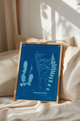 A framed botanical illustration of ferns and other plants is displayed on a bed, with a beige pillow and a window in the background.