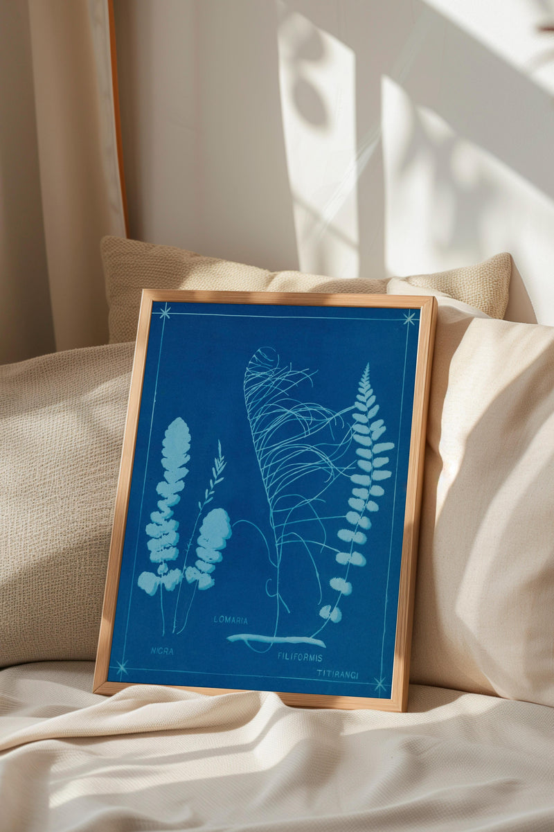 A framed botanical illustration of ferns and other plants is displayed on a bed, with a beige pillow and a window in the background.