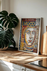 a framed mosaic artwork depicting a woman's face, placed on a wooden surface next to a potted plant.