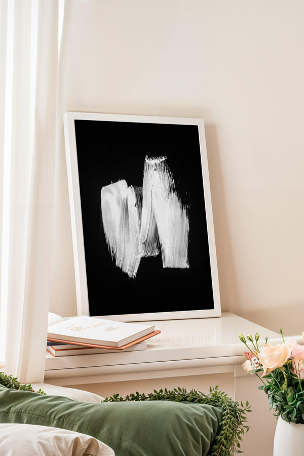 a framed black and white abstract painting on a white wall, with a white bookshelf below it. There is also a vase of flowers on the bookshelf.
