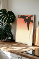 a framed artwork of two black birds in flight, displayed on a wooden surface next to a potted plant.