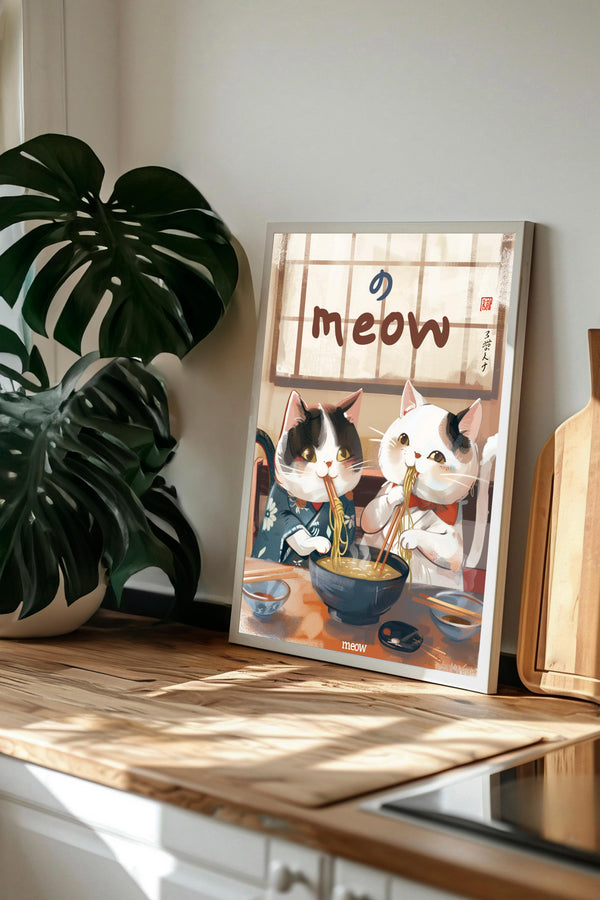A framed illustration of two cats sitting at a table, one of them eating noodles, is displayed on a wooden countertop next to a potted plant.