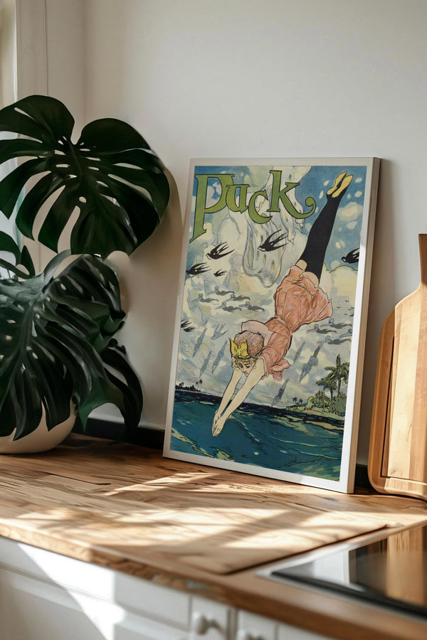 a framed vintage advertisement poster of a woman diving into the water, with the word "Puck" prominently displayed. The poster is placed on a wooden surface, with a potted plant and a wooden cutting board visible in the background.