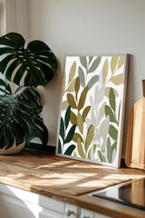 a framed print of a leafy plant design on a wooden surface, with a potted plant and a wooden cutting board visible in the background.
