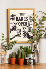 a framed poster with the text "The bar is open" and illustrations of people holding wine glasses and bottles, surrounded by potted plants and a camera on a wooden surface.