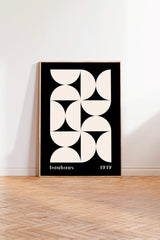 a minimalist black and white geometric poster with the text "bauhaus 1919" on it, standing on a wooden floor against a white wall.