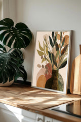 a framed print of a plant arrangement on a wooden surface, with a large monstera plant in the background.