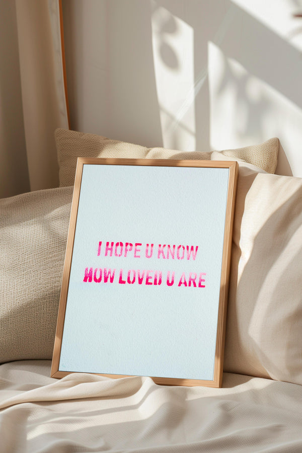 a framed sign with the text "I HOPE U KNOW HOW LOVED U ARE" displayed on it, placed on a bed with a beige pillow.