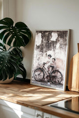 a framed photograph of two people riding a bicycle, with a potted plant and a wooden cutting board visible in the background.