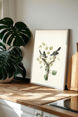 a framed print of a bird perched on a branch with leaves, placed on a wooden surface next to a potted plant.